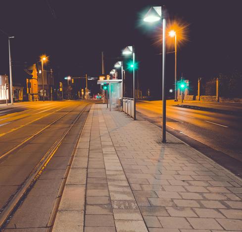 The image depicts a deserted urban street at night. There are tram tracks running alongside a paved sidewalk. Streetlights illuminate the area, and traffic lights can be seen ahead. There's a sheltered tram or bus stop visible on the right side of the tracks. The street appears quiet and empty.