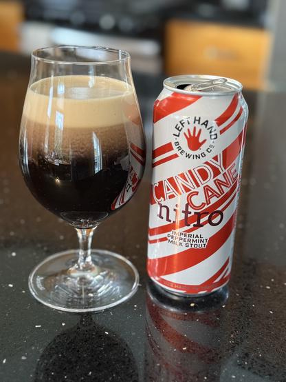 A tall snifter glass full of dark beer that is forming a thick tan head as nitro sheds into the dark brown liquid. The pint can is peppermint diagonal striped in white and red with the red left hand logo top center.