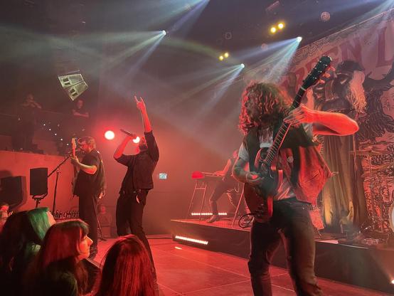 Green Lung performing on stage in a red light. There is the bassist, singer, keyboard player in the back and guitar player at the front. The singer is belting a line and doing the devil horns with his hand.