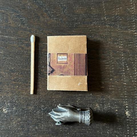 A matchstick, a tiny book titled "Saints," and a metallic hand-shaped object are arranged on a wooden surface.
