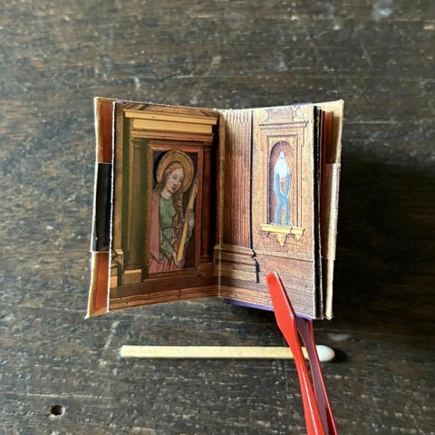 A pair of stamp tweezers holds a miniature book. The book open to a page spread featuring a collage of a photograph of a wooden cathedral model and illustrations. The backdrop is a close-up of two windows from wooden cathedral models. One window features an illustration of St. Agatha holding a candle, while the other displays an illustration of her attributes (pincers). A matchstick rests below the book for scale.