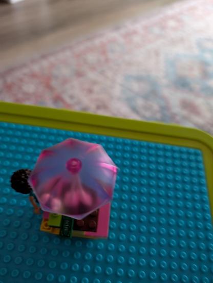 In a vibrant LEGO picnic scene, a mini doll with dark, curly hair sits comfortably on a yellow bench, part of a charming picnic table setup. She wears a light blue top, her tan skin tone adding warmth to the colorful display. Above her, a pink and translucent umbrella stands tall, casting a gentle shade over the table.
, 
, The picnic table itself is built on a pink base, creating a cheerful foundation for the scene. On the table, a small brown ice cream cone awaits, a delightful treat for the mini doll. Beside it, a green rectangular LEGO piece, representing money, adds a playful touch of realism.
, 
, The entire setup is placed on a larger blue LEGO baseplate, bordered by a bright green edge. This vivid contrast enhances the whimsical nature of the miniature world, where each brick—pink, yellow, green, brown, blue, and translucent pink—plays a part in crafting a lively and inviting picnic atmosphere.