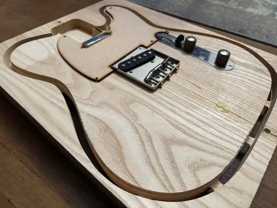 a telecaster body cut from ash and still attached to the extra wood from the blank with tabs.  the pickups, bridge, control cover, and a mock mdf pickguard have been put into place to visualize the final product.