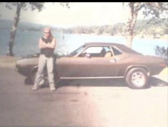 A man stands with crossed arms next to a vintage brown muscle car, parked by a scenic lake with trees in the background. The image has a vintage, faded quality.