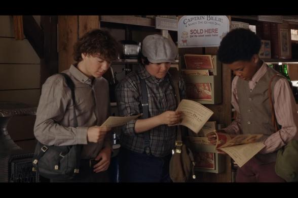 A screenshot from season 12, episode 2 of When Calls The Heart. There are 3 young boys standing in a mercantile shop, and each one is holding a comic book printed in newsprint in their hands, two of them are flipping through the pages. The middle one is leaning against a display rack that holds 3 different covers, and the sign at the top reads: "Captain Billie & Nancy The Genius Dog SOLD HERE, get the latest comic today"