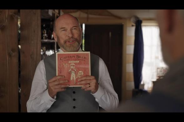 A screenshot from season 12, episode 2 of When Calls The Heart. The owner of a mercantile shop is standing in his shop, and holding up a comic book printed in newsprint in his hands. The cover is a faded red, and the title reads  "Captain Billie & Nancy The Genius Dog: The Great Escape". The cover shows a young blond girl wearing a space suit with a bubble helmet, running away from green martians, with her canine companion, a brown mutt wearing a space helmet and bandana, running alongside her.