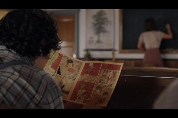 A screenshot from season 12, episode 2 of When Calls The Heart. We're looking over the shoulder of a young boy sitting in a schoolroom. As the teacher writes on the chalkboard at the front of the class in the background, he's holding a comic book open to read. The pages show the young girl lead character, and her canine companion, held captive by the martians.
