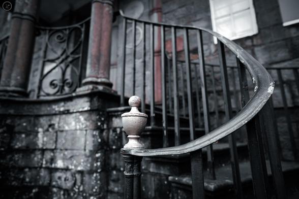 Shot of ornate railings up stairs and around pillared entrance to building, black paint peeling with rust