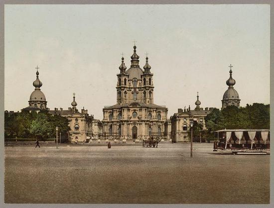 The image depicts an architectural structure that appears to be a cathedral or church with multiple domes and spires. The main building has a central dome flanked by four smaller domes, each topped with what seems like crosses. Surrounding this central edifice are additional buildings, including one on the left side featuring rounded arch windows and another larger complex in the background that complements the architectural style of the foreground structure.

In front of the church is an open plaza or courtyard paved with cobblestones. A few individuals can be seen walking across the area; however, their features are not clearly distinguishable due to the distance from which the photo was taken and its age. To the right side in the background, there's a horse-drawn carriage parked near what appears to be an outdoor café or restaurant with covered seating.

The overall atmosphere of the image suggests it is historical, possibly dating back to late 19th century Russia based on architectural styles and urban elements present at that time. The sepia tone indicates that this photo might have been taken during a period when color photography was not widely used for everyday images or events; instead, these types of photographs were often created using the Photochrom process which produced vibrant prints from glass negatives.

The image is labeled with text indicating its source and context: "St. Pétersbourg," meaning Saint Pe [...]