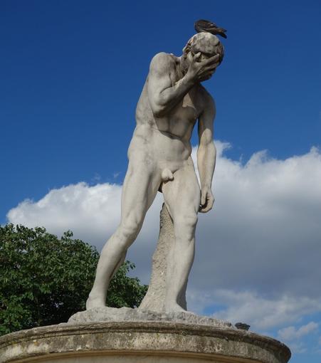 A frontal view of a light grey stone statue of a nude male figure with his head bowed and his hands covering his face in desperation. He stands on a round, tiered pedestal, with a tuft of green foliage visible on the left side of the frame. A pigeon is perched on the statue's head. The background is a bright blue sky with fluffy white clouds.