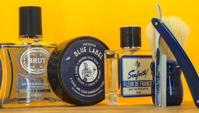 (Le Chat Mistral description)
This image displays a collection of men's grooming items against a bright yellow background. On the left, there's a glass bottle of Brut aftershave with a black cap. Next to it is a round black container of Razorock Blue Label shaving soap. In the centre, there's a small bottle of Seaforth Fleur de France eau de toilette. To the right, there's a straight razor with a blue handle and a shaving brush with a matching blue handle. The items are neatly arranged, emphasising classic shaving and fragrance products.