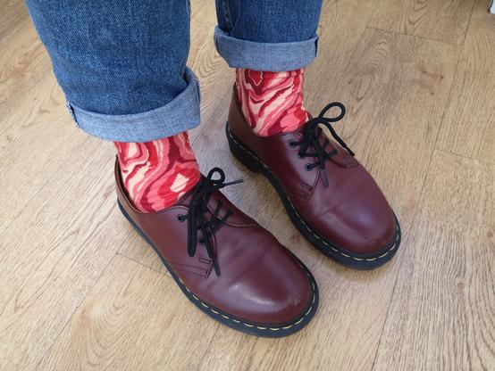 Photo de 2 jambes en jean bleu ourelé et chaussures DocMartens bordeaux. Entre les deux on voit des chaussettes avec des motifs en formes de vagues irrégulières dans des tons rouge, rose, beige, orange