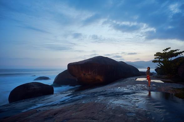 A rocky shore is seen, with some larger rocks or boulders in the back towards the shoreline. In the middle is a larger rock in form of a shark. A woman is standing to the right looking at the sea. From the right comes a small river that runs towards the sea. The rocks and the ground are of a reddish color with grey patches. The sea looks milky like mist due to the long exposure. The sky is still of a light blue though the sun has already set.