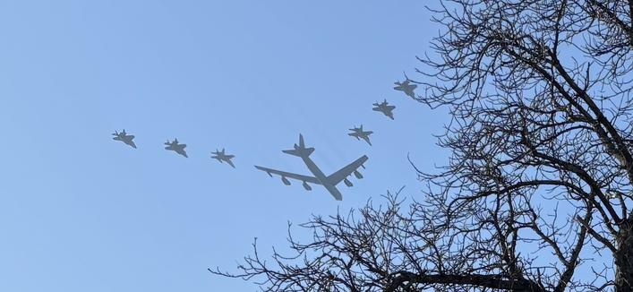A formation of military aircraft flying in the sky, including a large plane trailing several smaller fighter jets. The silhouettes are set against a clear blue background, with bare tree branches visible in the foreground.