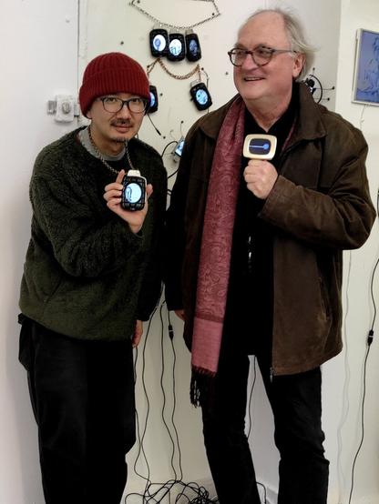 Reinhard Hampel and Nao Tanaka wearing Vijou : Reinhard his Golden TV Vijou, a golden jewel shaped like a 1970s TV, Nao a Grenade Vijou shaped like a hand grenade. They stand in front of the Sigrid Coggins exhibit in Galerie Satellite composed of Grenade Vijou and Digital Bug.