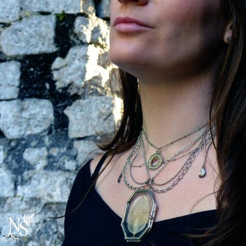 English A woman's neckline with a black top and a multichains silver necklace that has tiny moon crescents on the sides, a moon phases pendant and a glass medallion showcasing a real lunaria annua disc that looks like the moon. At the back we can see a very old stone wall covered in moss and the sunlight breaking through.
•
Français Un décolleté de femme avec un haut noir et un collier en argent multi-chaînes avec de petits croissants de lune sur les côtés, un pendentif des phases de la lune et un médaillon en verre contenant un véritable disque de lunaria annua qui ressemble à la lune. À l'arrière, on peut voir un très vieux mur de pierre recouvert de mousse et la lumière du soleil qui passe à travers.