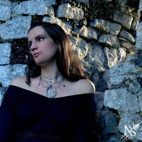 English A young woman standing next to a very old stone wall covered in moss and the sunlight breaking through a hole in it. She wears a black lace dress and long dark blonde hair. Her necklace is an outstanding piece made of silver multichains, with tiny moon crescents on the sides, a moon phases pendant and a glass medallion showcasing a real lunaria annua disc that looks like the moon.
•
Français Une jeune femme se tient à côté d'un très vieux mur de pierre couvert de mousse et la lumière du soleil passe à travers un trou dans le mur. Elle porte une robe noire en dentelle et de longs cheveux blonds foncés. Son collier est une pièce exceptionnelle faite de chaînes multiples en argent, avec de minuscules croissants de lune sur les côtés, un pendentif des phases de la lune et un médaillon en verre mettant en valeur un véritable disque de lunaria annua qui ressemble à la lune.