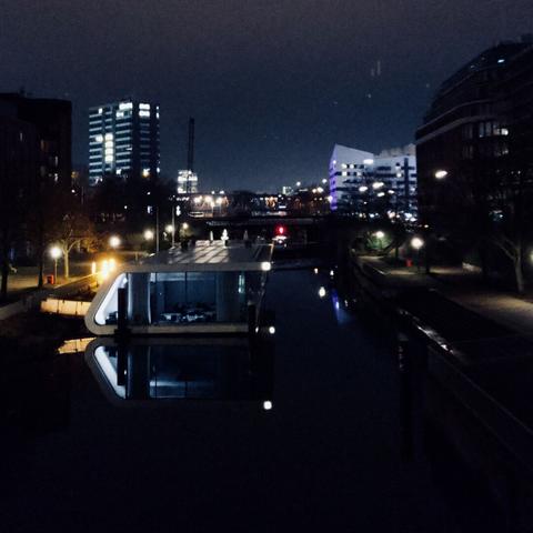Houseboats on a canal in between office buildings. A concrete desert with straight waterways. In the background is a train bridge.