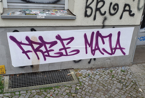 Bild eines Graffiti. weißer Streifen auf einer grauen Hauswand (unter einem mit Stickern beklebten Fensterrahmen) auf welchem in lila Großbuchstaben. FREE MAJA steht.