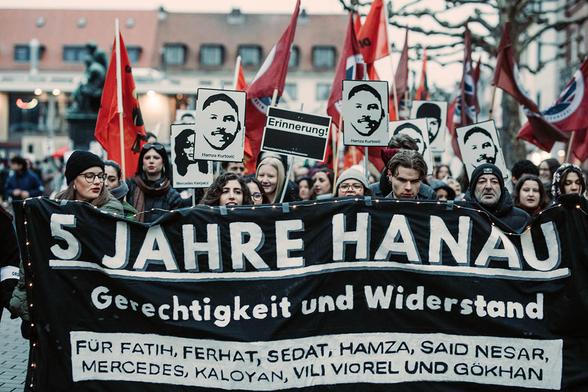Das Foto zeigt das Transparent mit dem Text: "5 Jahre Hanau - Gerechtigkeit und Widerstand - Für Ferhat Unvar, Hamza Kurtović, Said Nesar Hashemi, Vili Viorel Păun, Mercedes Kierpacz, Kaloyan Velkov, Fatih Saraçoğlu, Sedat Gürbüz und Gökhan Gültekin". Dahinter zahlreiche Menschen mit Fahnen und Tafeln mit den Gesichtern und Namen der Opfer.