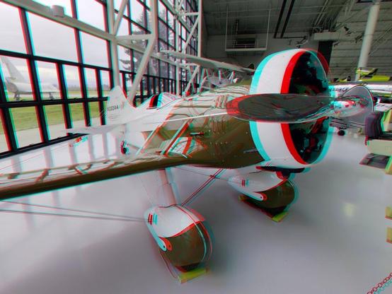 Inside an air museum, a beautiful, bulbous, red and white 1930s racing monoplane with a chrome propeller. Stereoscopic, color view for anaglyph (red/blue) glasses.