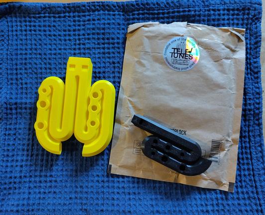 On a blue cloth, there's a yellow plastic flute with six holes in the shape of two snails and next to it a brown envelope. On the envelope, there is a black plastic flute with six holes in the shape of a snail.