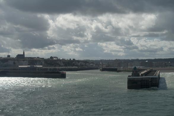 Photograph of the seafront of Dieppe seen from a departing ferry. The surface of the sea is green and rugged, slightly swirly.
The sky, taking nearly half of the picture, has large clouds but also many bright patches of sunlight.
The city itself is divided by the port in a left half with cliffs and a church on top and an urban right half with a beach and the seafront buildings.
A long concrete pier extends towards the viewer from the port.