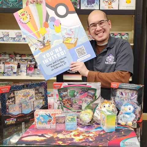 Man wearing a hairy tarantula branded collared t-shirt, holding up a poster saying "You're invited! Come and Celebrate Pokemon Day on February 27th" there are two hot air baloons, one shaped like a pokeball carrying various pokemon on the poster. 

There is also a bunch of assorted pokemon card game merch on a table and on the shelves around them.