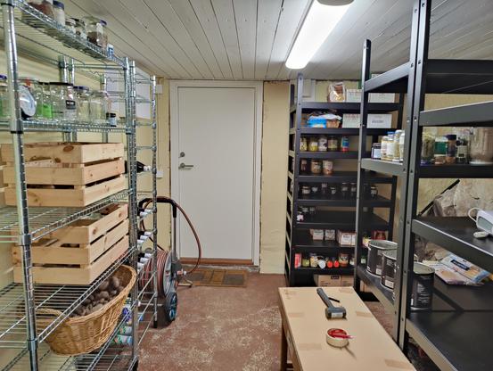 A brightly lit basement with metal shelving units. On the shelves are crates and baskets with potatoes and jars with various jams, sauces, spices, fruits, and vegetables.