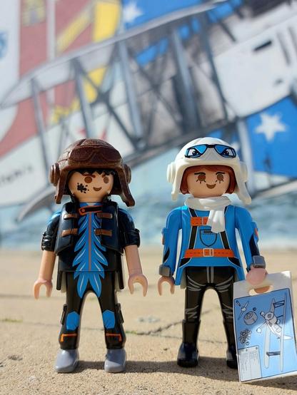Bonecos aviadores playmobil, vestidos de azul, o homem com capacete antigo de aviador castanho e a menina com capacete antigo  de aviador branco, frente a um mural com avião bimotor e bandeiras de Portugal e Brasil