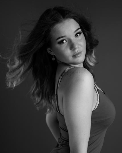 Black and white studio portrait of a young woman with wavy hair, wearing a fitted top. She gazes at the camera with confidence, her expression highlighting elegance and style.