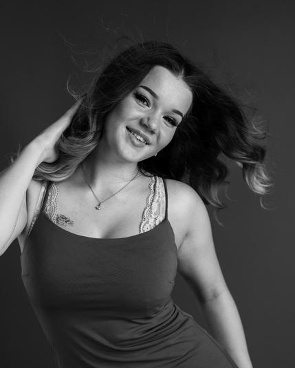The image is a black-and-white portrait of a smiling person with shoulder-length hair. They are wearing a sleeveless top with lace and appear to be in a playful or relaxed pose, gently holding one hand to their hair.