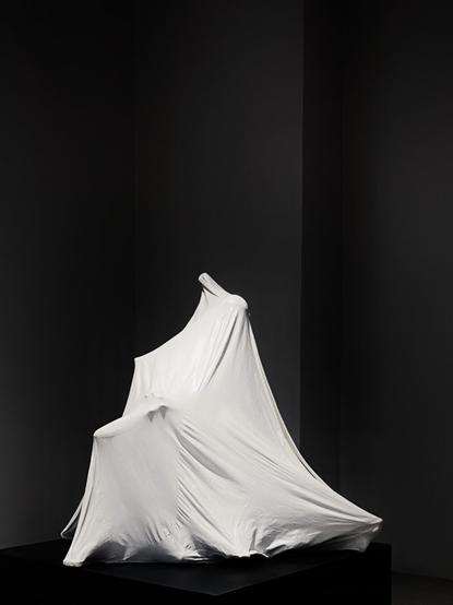 An image of a sculpture depicting a figure underneath a white sheet mounted on a box against a dimly lit display wall.