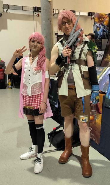 Two cosplayers in front of an art booth. They have similar pink hair. Serah wears a pink gown, a white blouse, a red-white checkered skirt and black overknee socks. Lightning wears a white coat with brown applications, a brown sweatshirt and brown short pants and boots.
