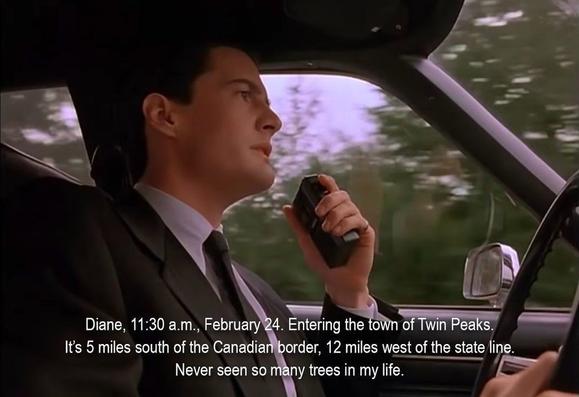 A man in a suit, driving a car and speaking into a tape recorder. He is saying, "Diane, 11:30 a.m., February 24. Entering the town of Twin Peaks.  It’s 5 miles south of the Canadian border, 12 miles west of the state line.  Never seen so many trees in my life. "