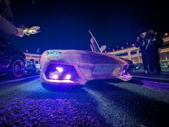 Front low shot of a Lamborghini murcielago completely covered in tiny white and pink crystals, showing a pink led fog light and a Japanese license plate "1"