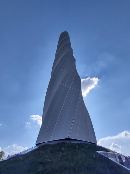 Das Bild zeigt einen modernen, spiralförmig gedrehten Turm, der imposant in den Himmel ragt. Die Fassade des Turms besteht aus glatten, metallischen Platten mit feinen Linien, die die Drehbewegung der Struktur betonen. Der Turm steht auf einem begrünten Hügel, dessen sanfte Erhebung ihn noch eindrucksvoller wirken lässt. Der Himmel im Hintergrund ist blau mit einigen verstreuten, weißen Wolken. Die Aufnahme wurde aus einer niedrigen Perspektive gemacht, wodurch der Turm besonders hoch und monumental erscheint. Die moderne Architektur und das innovative Design lassen darauf schließen, dass es sich um ein technisches oder wissenschaftliches Bauwerk handelt.