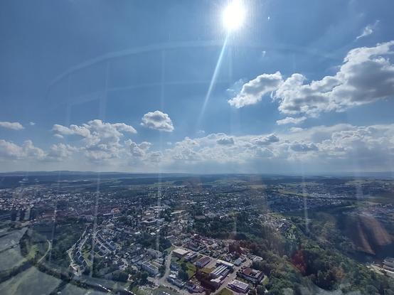 Das Bild zeigt eine weitläufige Landschaft mit einer Stadt, die sich über ein großes Gebiet erstreckt. Die Perspektive ist hoch gelegen, aus einem hohen Aussichtsturm aufgenommen. Der Himmel ist blau mit einigen weißen Wolken, und die Sonne strahlt hell, wobei ihr Lichtstrahl deutlich zu sehen ist. Im Vordergrund sind verschiedene Gebäude, Wohnhäuser und Straßen zu erkennen, die sich durch die Stadt schlängeln. Im Hintergrund erstreckt sich eine ländliche Landschaft mit grünen Feldern und Wäldern. Die Reflexionen im Glas deuten darauf hin, dass das Foto durch eine Fensterscheibe aufgenommen wurde.