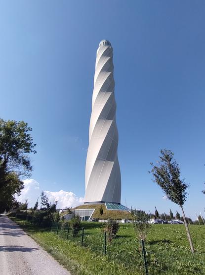Das Bild zeigt einen hohen, modernen Turm mit einer markanten, spiralförmigen Fassade. Der Turm steht frei in einer grünen Umgebung mit einer umzäunten Wiese, kleineren Bäumen und einem Schotterweg, der an ihm vorbeiführt. Am unteren Bereich des Turms befindet sich ein Gebäude mit begrüntem Dach und großen Glasflächen. Der Himmel ist klar und blau, mit wenigen Wolken am Horizont. Die Perspektive des Bildes betont die Höhe des Turms, der majestätisch in den Himmel ragt. Im Hintergrund sind einige geparkte Autos zu sehen