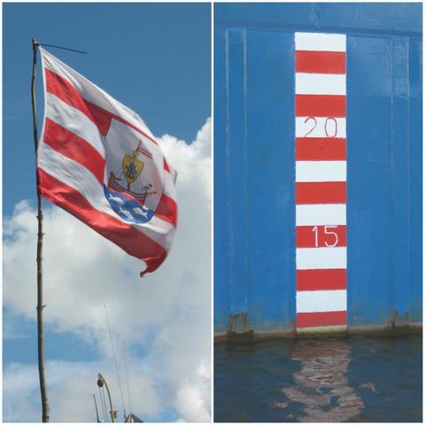 Fotocollage van 2 staande beelden.
1 een wapperende vlag voor een blauwe lucht met witte wolken. De vlag heeft wit-blauw-witte strepen met in het midden een afbeelding van een vikingschip.
2 op de blauwe zijwand van een schip een smalle baan met wit-rood-witte strepen en enkele getallen (15 en 20)