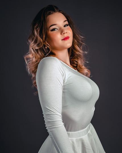 A young woman with wavy brown hair and subtle makeup posing elegantly in a studio setting, wearing a fitted white long-sleeve top and skirt, against a dark background.