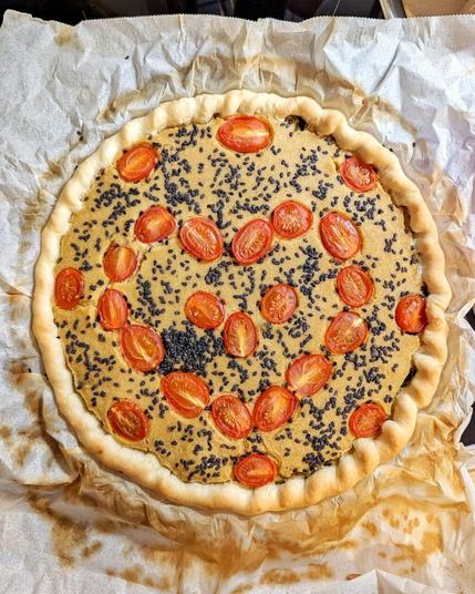 Vogelblick auf eine Backform mit einer Quiche darin. Backpapier, leicht gebräunt, wallt über den Rand der Form und bildet optisch einen Rahmen. Der Teig der Quiche ist leicht gebräunt, die Füllung besteht aus einem fest gewordenen, gelb-beigen Guss. Darauf ist schwarzer Sesam gestreut und halbierte in den Teig gedrückte und mitgebackene Kirschtomaten bildern ein großes, rotes Herz.