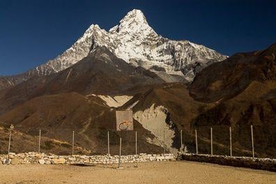 A colour photograph. There is a backboard and fenced basketball court in the foreground. There is a deep valley behind it and further, above there is a snow capped, sharp shape, high mountain.