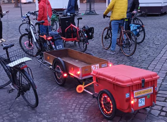 Auf dem Kopfsteinpflaster des Domplatzes von Münster stehen Fahrräder unter anderem ein Lastenrad beladen mit einem Lautsprecher und ein Fahrradlastenanhängergespann mit 2 Anhängern. Am 2 Anhänger ein Schild "Long Vehikel".