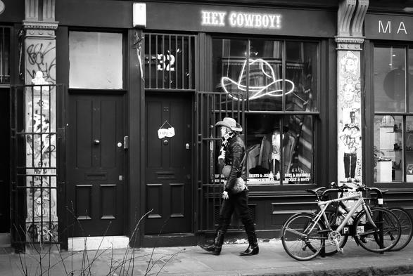 Black and white photo of a shop called Hey Cowboy! in Cheshire Street, just off London's famous Brick Lane. Just outside the shop, about to go in, is a man in a Stetson cowboy hat and mid-calf length shiny black boots with no spurs. He is also wearing a black quilted leather-look jacket and a light-coloured furry bag on a strap over one shoulder. So probably not a real cowboy, then.