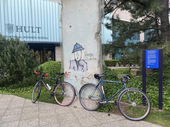 Two bicycles are propped up in front of a piece of the Berlin Wall, now located in front of Hult International Business School in Boston, MA, USA. The piece of the wall is protected by glass barriers, and the graffiti on the wall depicts a person with a blue hat, with partial texts reading "Quick (2 min", "ME", and "FILA".

It is summer and there are green grasses, bushes, and trees also in the frame.

A sign with blue background and white lettering describes the wall piece: "On November 9, 1989 the citizens of both East and West Berlin joined together to destroy the cement barrier that had separated them since 1961. This small section of the Wall was once located near Bernauer Strasse, a street famously bisected by the border. The side emblazoned with graffiti faced West Berlin and the bare side, East Berlin. EF Education First dedicates this important piece of history to those throughout the world who share our commitment to opening the world through education. September 25, 2000 EF"