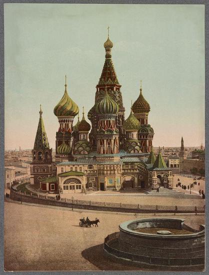 The image displays a detailed colored postcard showing the iconic St. Basil's Cathedral, located in Red Square in Moscow, Russia. The cathedral is characterized by its vivid green onion-shaped domes with golden tips and intricate architectural details. Surrounding the main structure are smaller buildings that complement its grandeur, including what appears to be an archway entrance on one side.

In front of St. Basil's Cathedral, a small circular pond or fountain can be seen at the forefront, adding to the overall urban scene depicted in the postcard. The street leading up to and surrounding Red Square is populated with pedestrians and horse-drawn carriages indicative of the late 19th-century setting.

The color palette consists primarily of greens from the domes, warm reds on some buildings' facades, earth tones for other structures and pathways, and a muted sky tone in the background. The postcard's vintage quality is suggested by its artistic rendering style rather than photographic realism, capturing an early 20th-century view of Moscow's most famous landmark.

In terms of additional information about this image, it may be linked to "Orthodox churches" as St. Basil's Cathedral serves a religious function in the Orthodox tradition and was built between 1555-61 during Ivan the Terrible’s reign. The photograph could also relate to Russia (Federation) Moscow specifically due to its central location within the city, being [...]