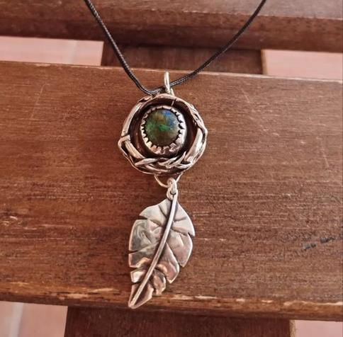 a pendant with black cord. it has a silver leaf and a silver circle with a blue and green gemstone in it. it's placed on brown wood