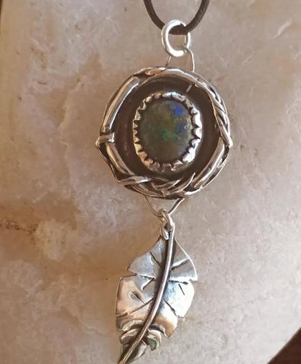 a pendant with black cord. it has a silver leaf and a silver circle with a blue and green gemstone in it. it's placed on white pebble
