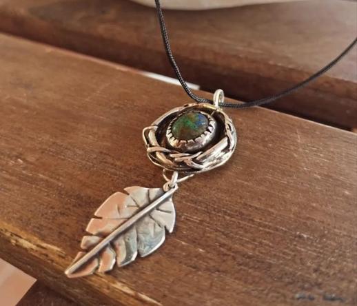 a pendant with black cord. it has a silver leaf and a silver circle with a blue and green gemstone in it.